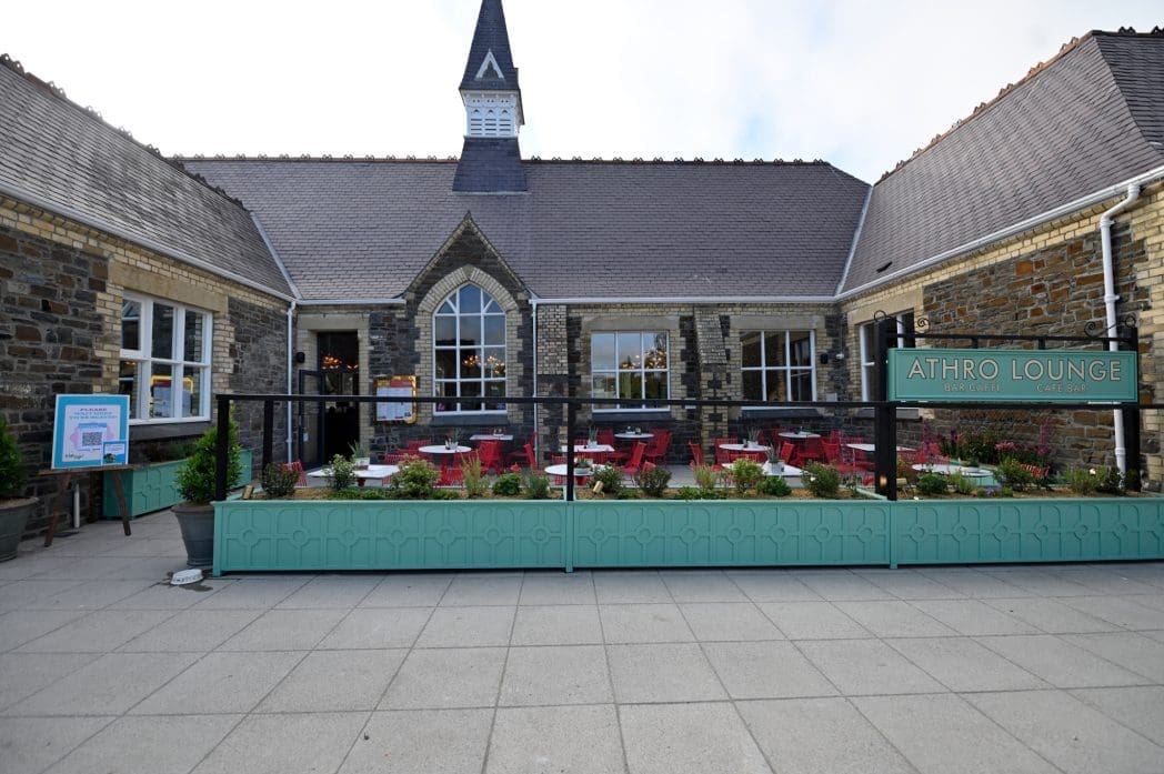 A stone building with large windows and a pointed tower, Athro Lounge features an inviting outdoor seating area with red chairs and tables. A sign reads "ATHRO LOUNGE," and lush plants border the patio.