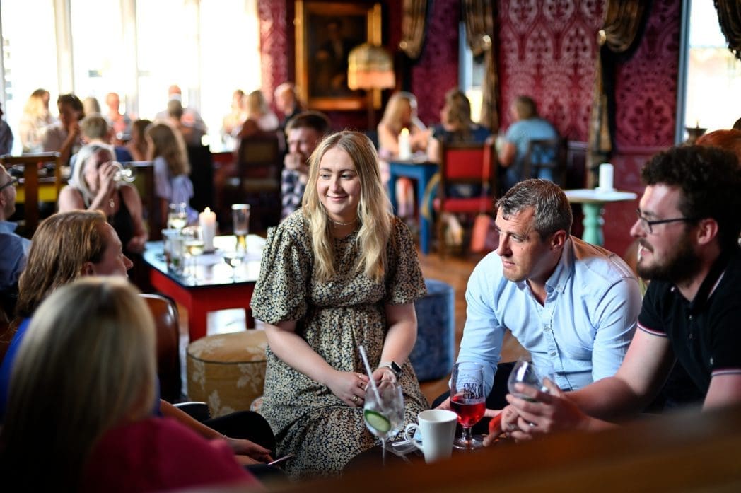 A group of people sit together in a warmly lit, busy Athro restaurant, chatting and smiling. The atmosphere is lively, with drinks on the table and other guests in the background.