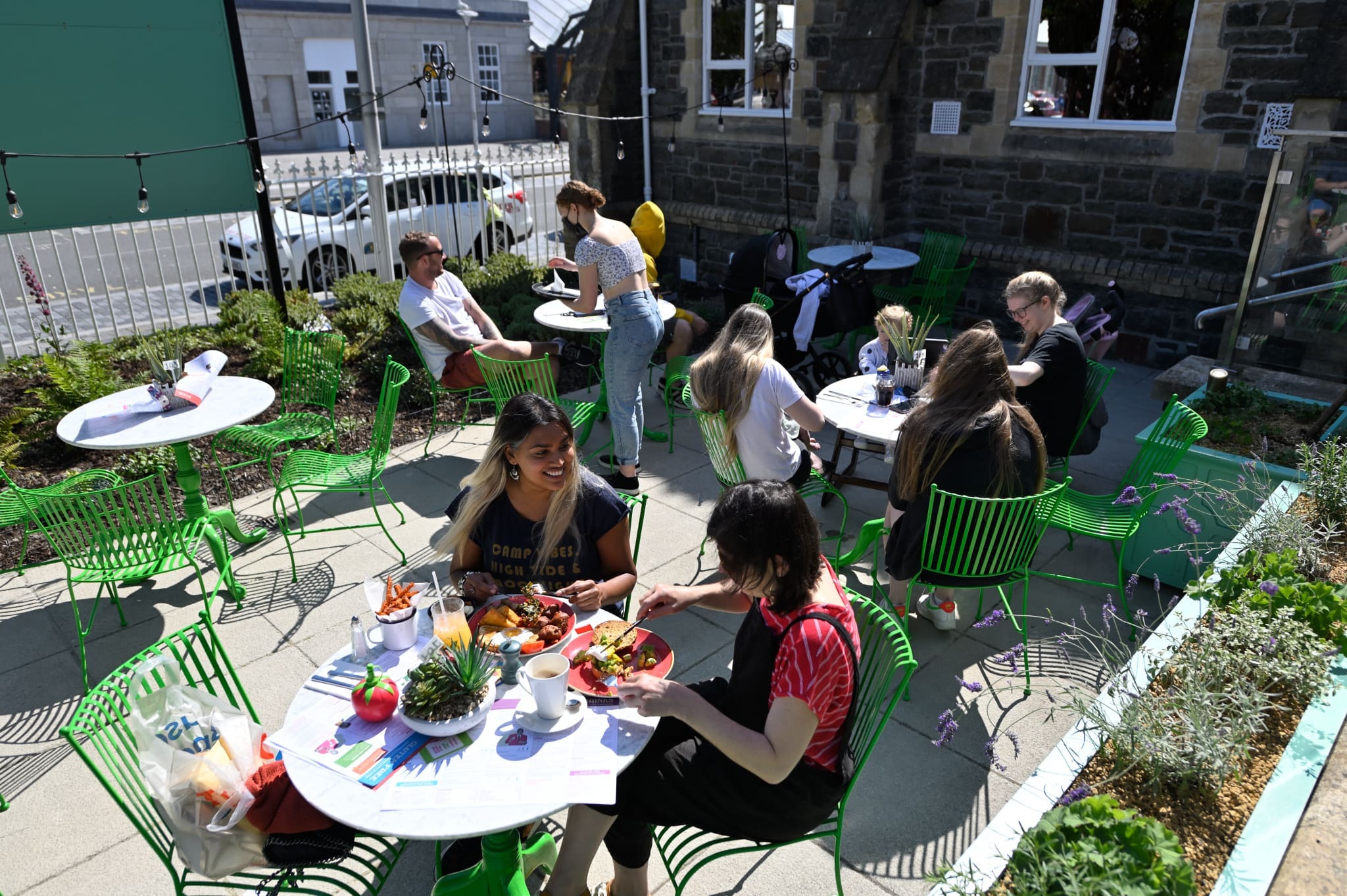 People sitting at green tables and chairs outdoors, enjoying food and drinks in a sunny café garden. Some are chatting and laughing with athro friends, while others are relaxing. The area is decorated with flowers and string lights.
