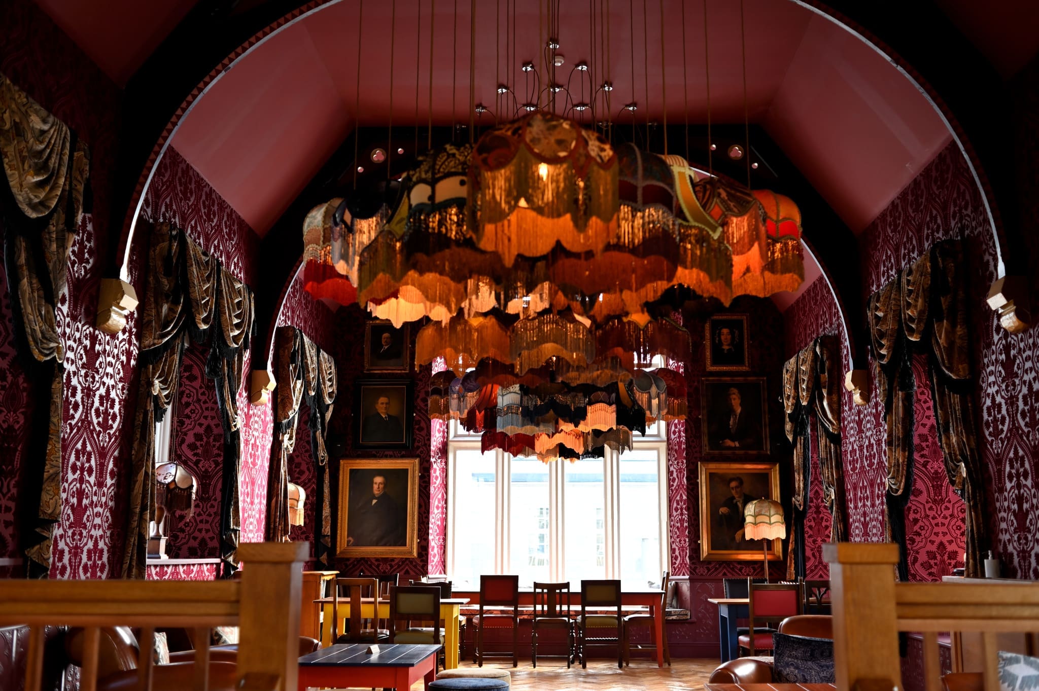 A lavish Athro room with ornate red wallpaper, vintage lampshades hanging from the ceiling, large windows in the back, and framed portraits on the walls. Heavy drapes and classic furniture add to the luxurious atmosphere.