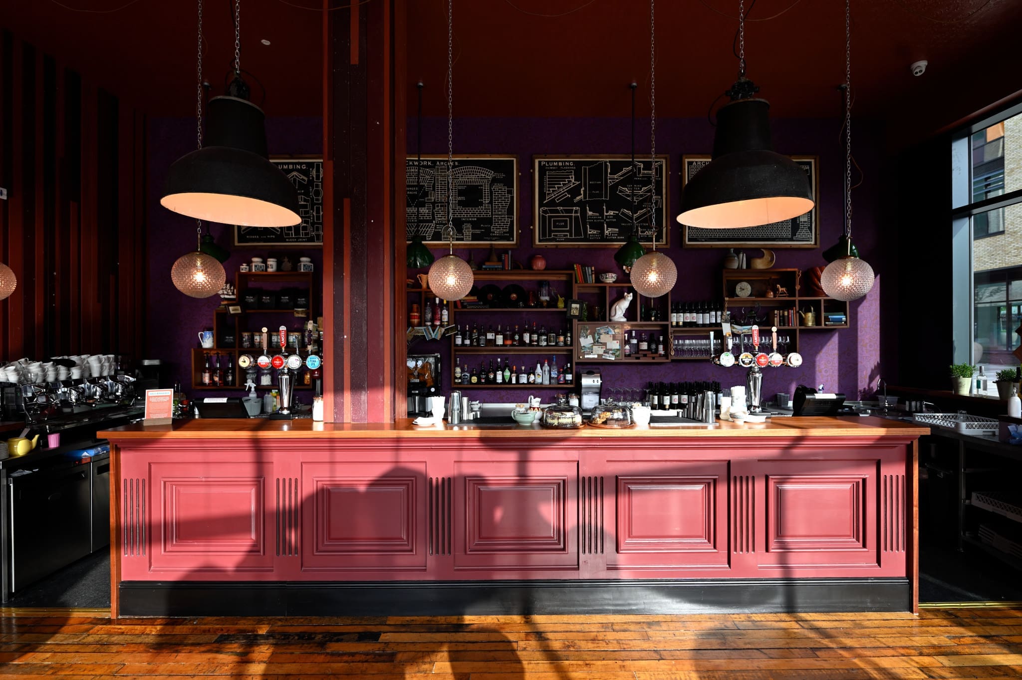 A stylish bar with a burgundy counter, hanging lights, shelves stocked with bottles, and art—featuring a playful Gatto—on the purple wall. Sunlight casts shadows on the wooden floor and bar front.