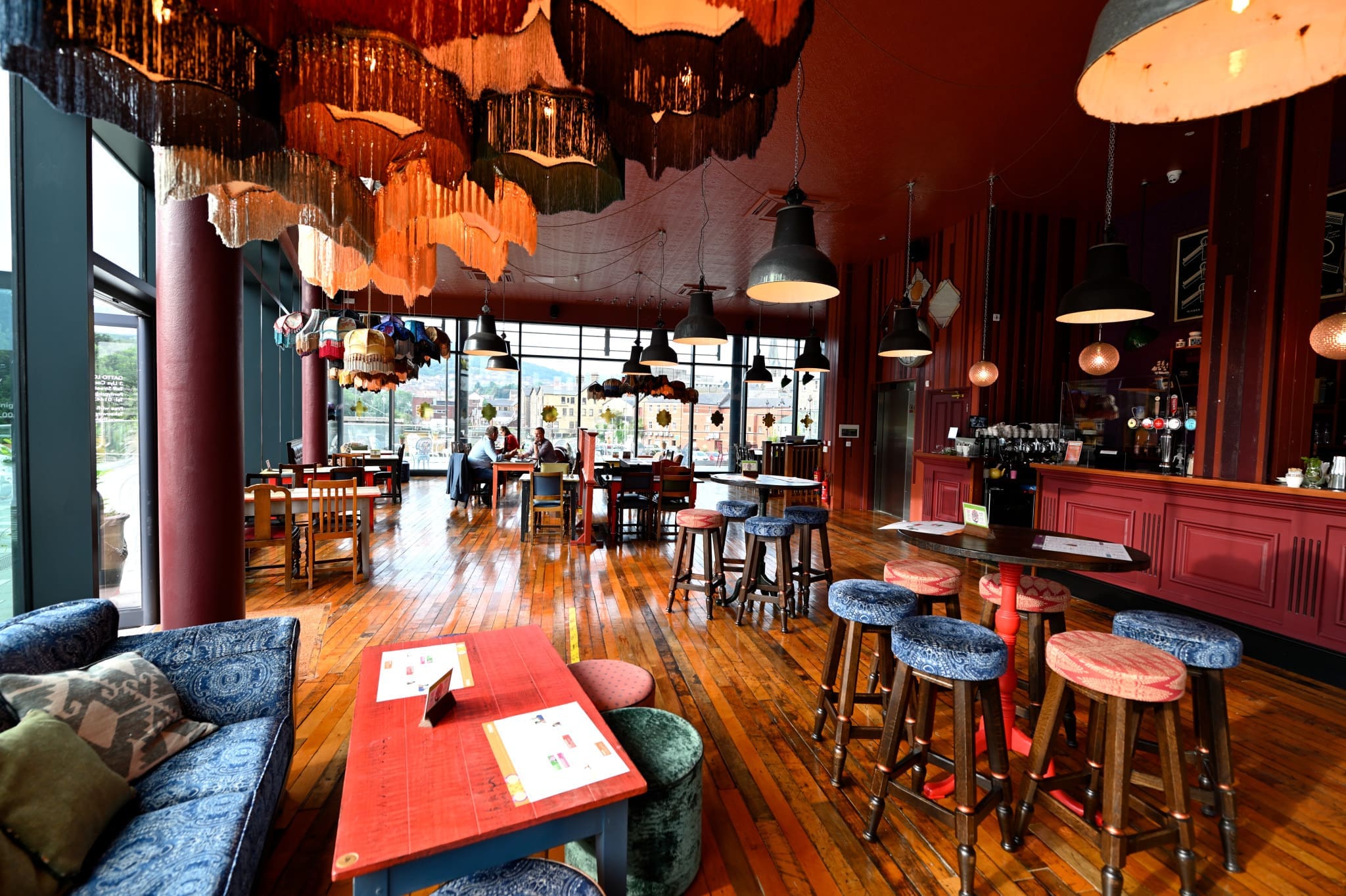 A vibrant restaurant interior with colorful hanging lamps, eclectic furniture, wooden floors, and large windows letting in natural light. The inviting Gatto bar is visible on the right, with tables and chairs arranged spaciously throughout.