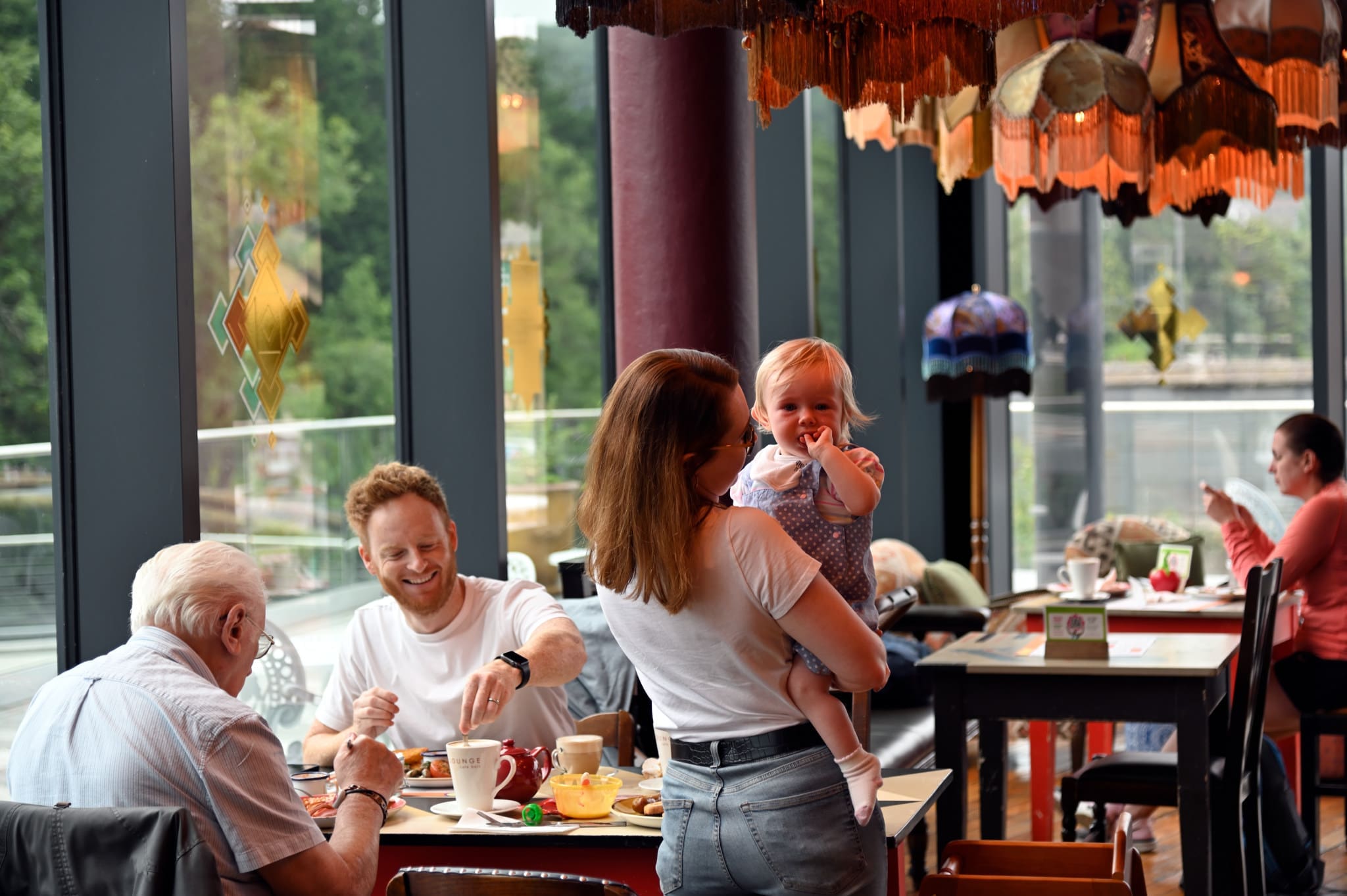A family enjoys breakfast at a restaurant with large windows. An older man and a younger man sit at the table, while a woman stands holding a smiling toddler and a plush Gatto toy. Other diners are visible in the background.