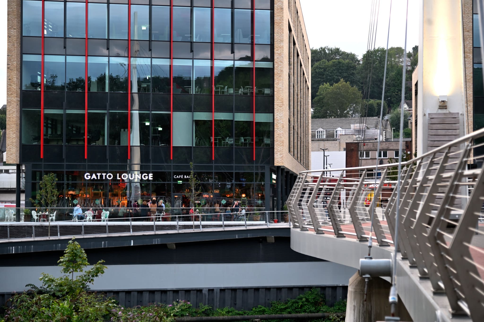 A modern building with large glass windows houses Gatto Lounge cafe bar. People are visible inside Gatto, while a footbridge with metal railings crosses in the foreground. Trees and more buildings appear in the background.