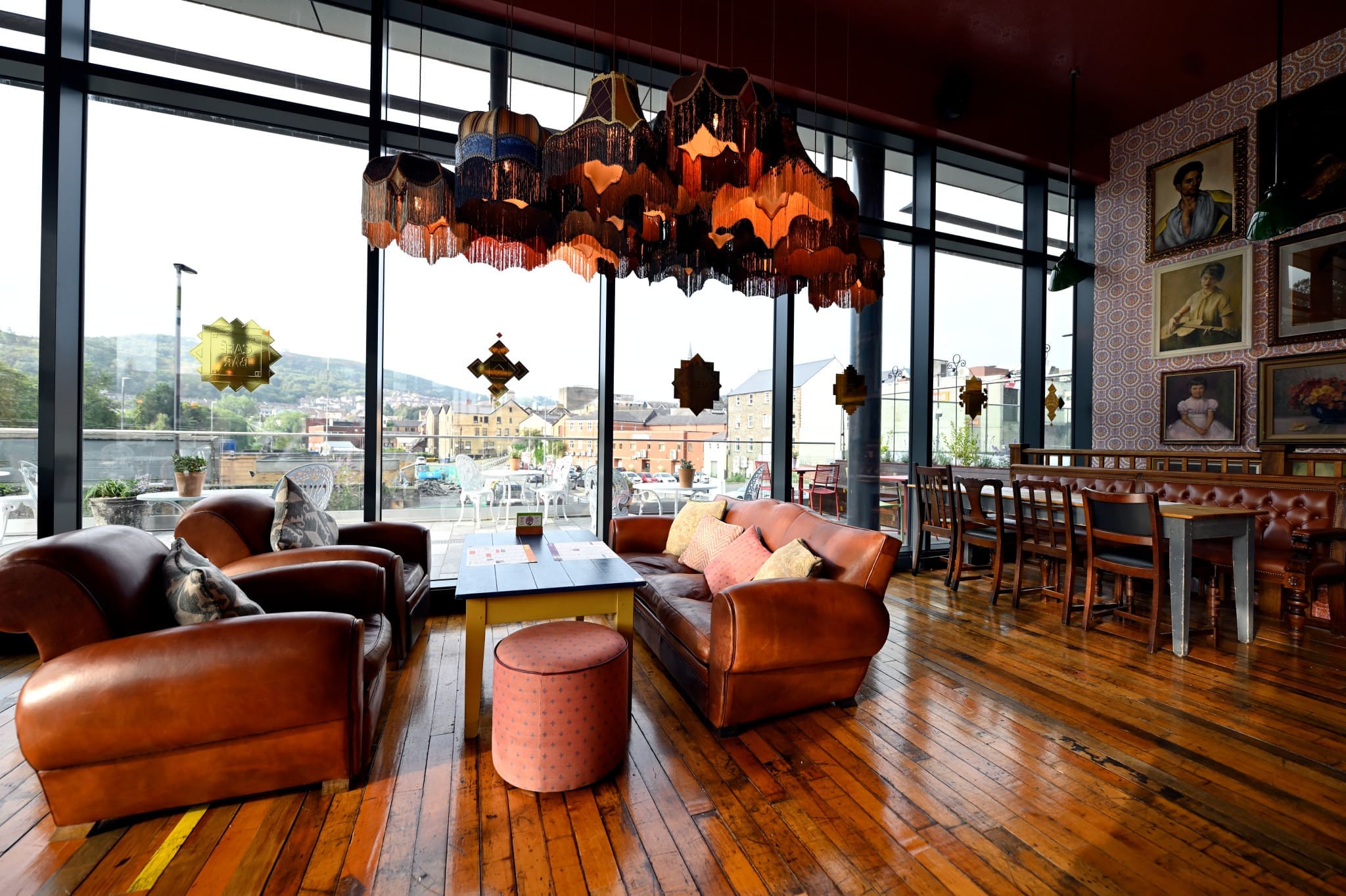 A cozy lounge with leather sofas, colorful cushions, eclectic hanging lamps, wooden floors, large Gatto windows with a city view, and art-covered walls.