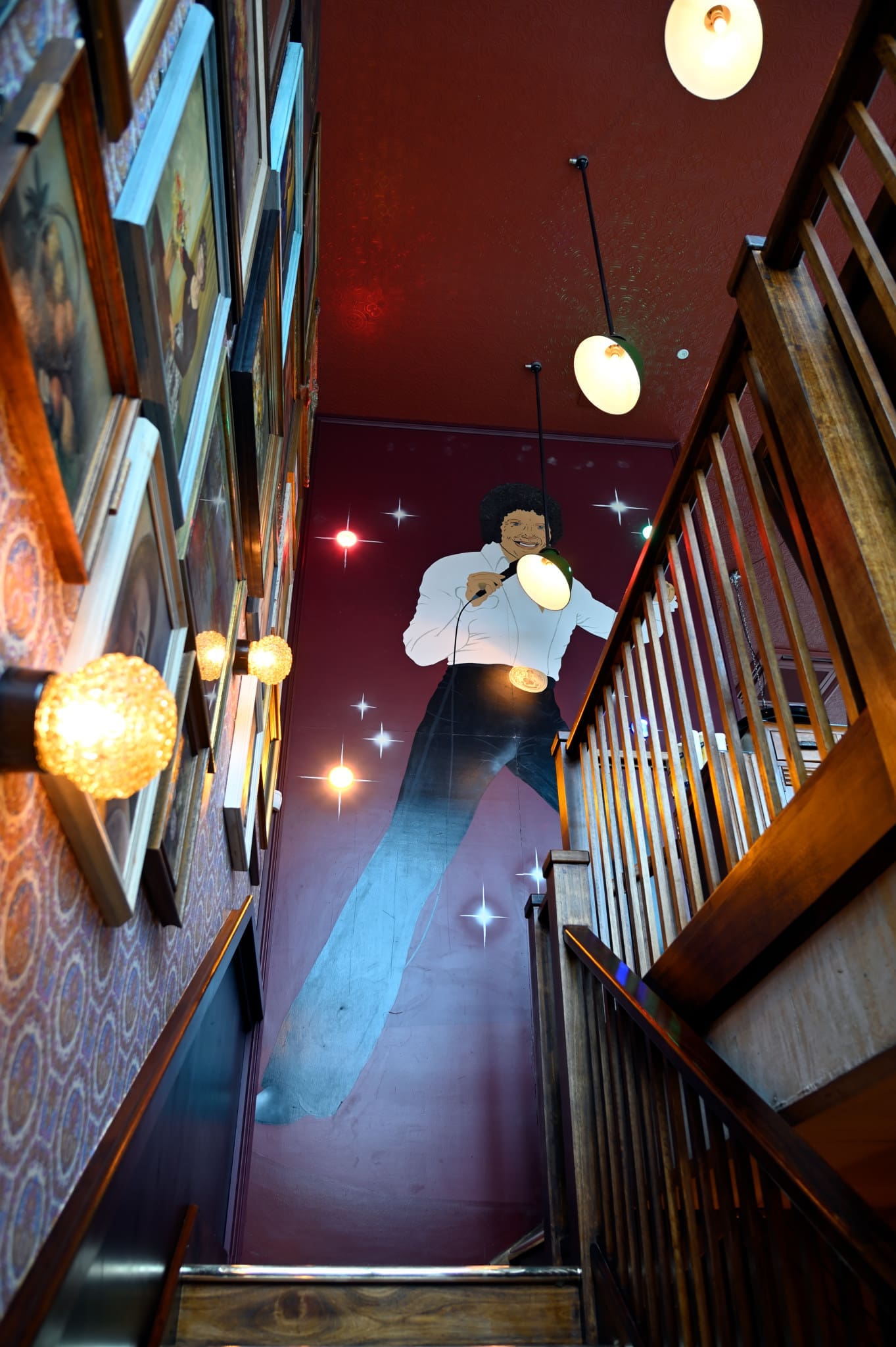 A staircase with wooden railings leads up to a wall featuring a large mural of a singer holding a microphone, surrounded by colorful lights, while framed pictures and a playful Gatto accent the adjacent wall.