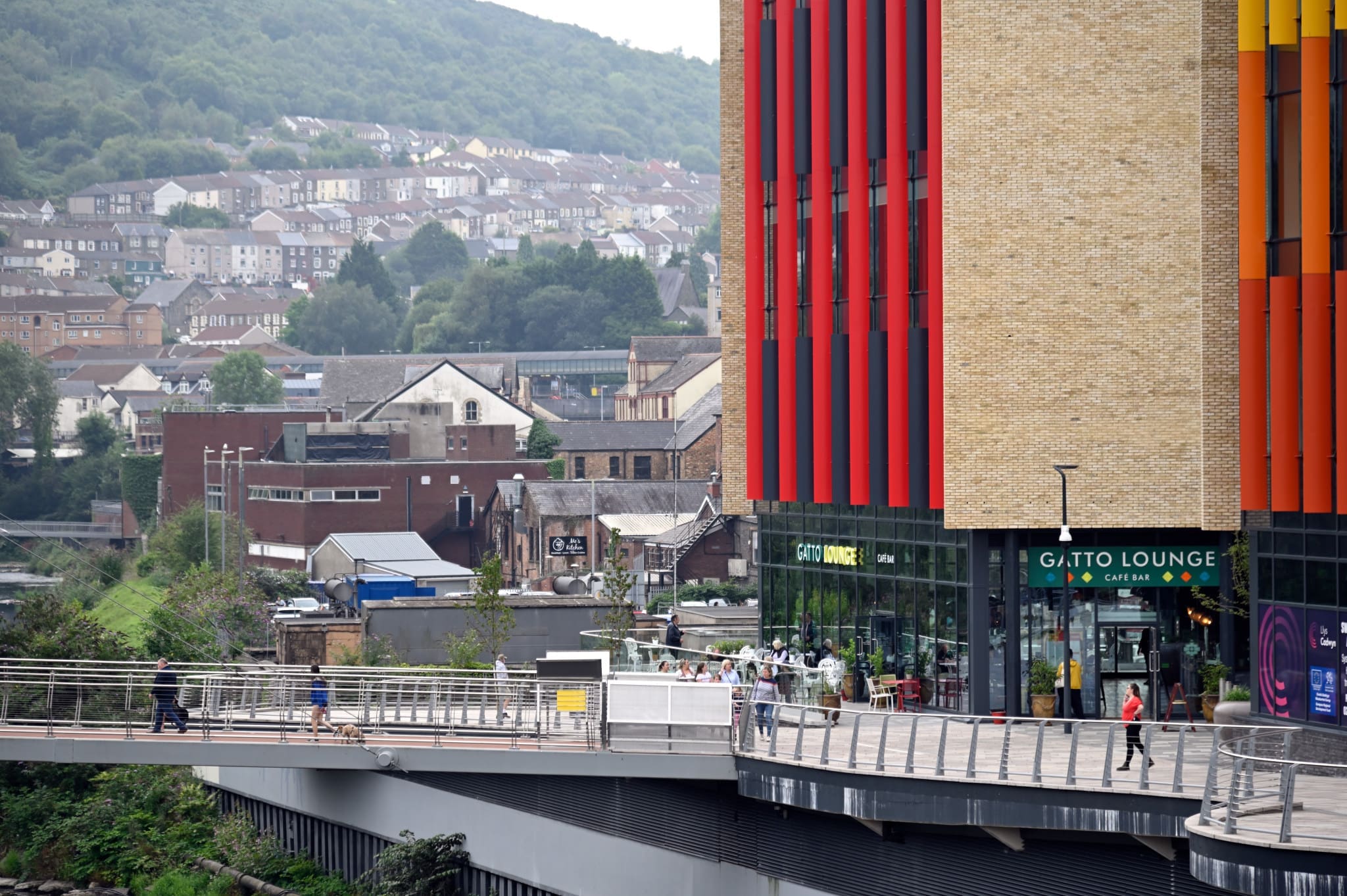 A modern building with vertical red and orange panels stands near a bridge, where people—and the occasional curious gatto—walk outside. Behind it, a hillside is covered with houses and trees under a cloudy sky.