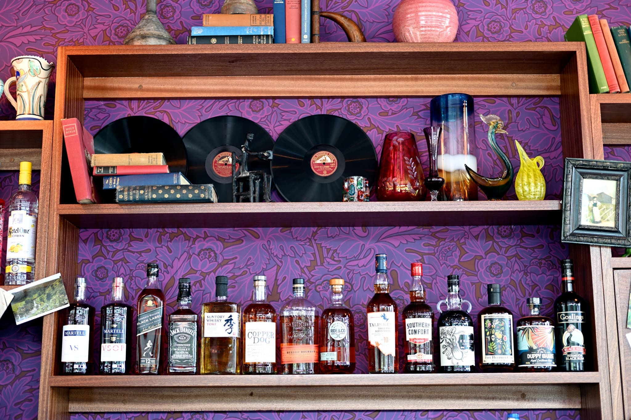 Wooden shelves against a purple floral wall hold vinyl records, books, decorative items, and framed photos—including one featuring a playful Gatto. The lower shelf displays an assortment of liquor bottles in various shapes and sizes.