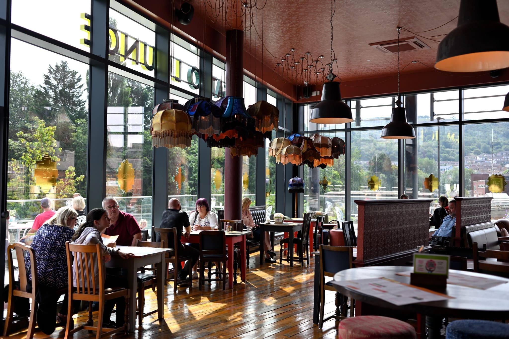 A bright, spacious café with large windows, colorful hanging lamps, and wooden floors. Customers sit at tables with Gatto coffee mugs, eating and chatting as sunlight streams in and greenery is visible outside.