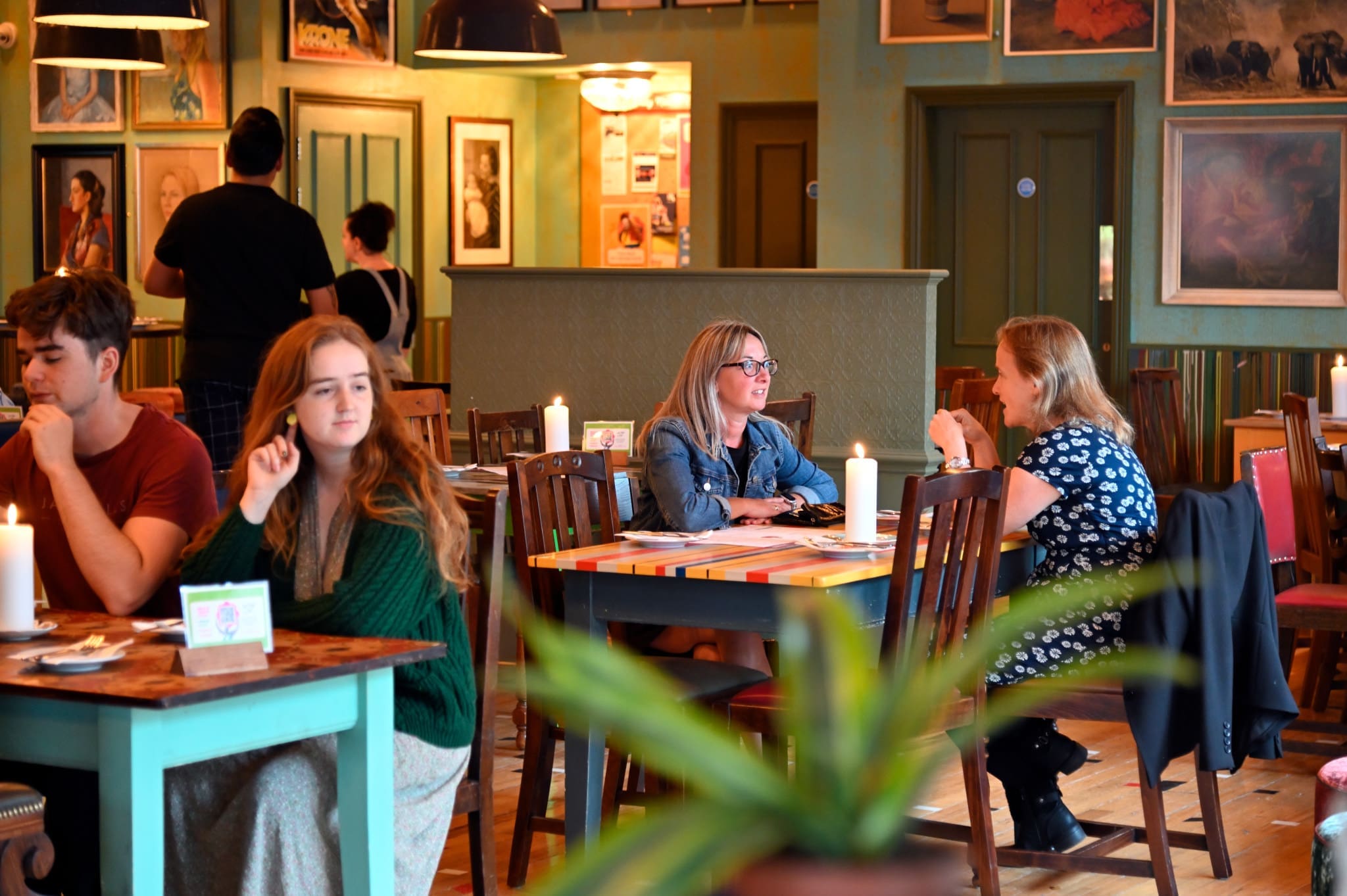 People sit at tables in Vito, a cozy, warmly lit restaurant, engaged in conversation. Some diners are talking while others look around. Art and photographs decorate the walls, with a green plant visible in the foreground.