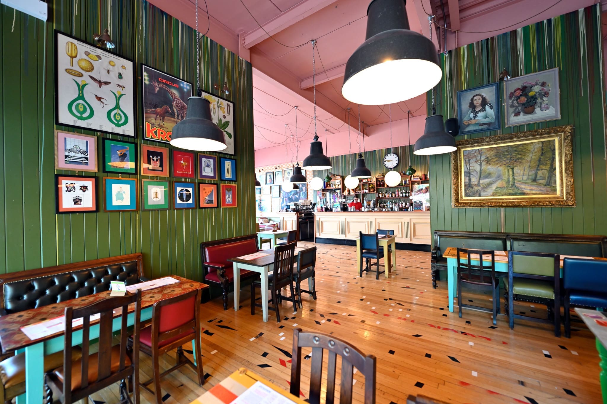 A colorful café with eclectic wall art, vintage posters, and framed pictures. Wooden tables and chairs fill the polished floor. Large black pendant lights hang from the pink ceiling above Vito’s inviting seating area.