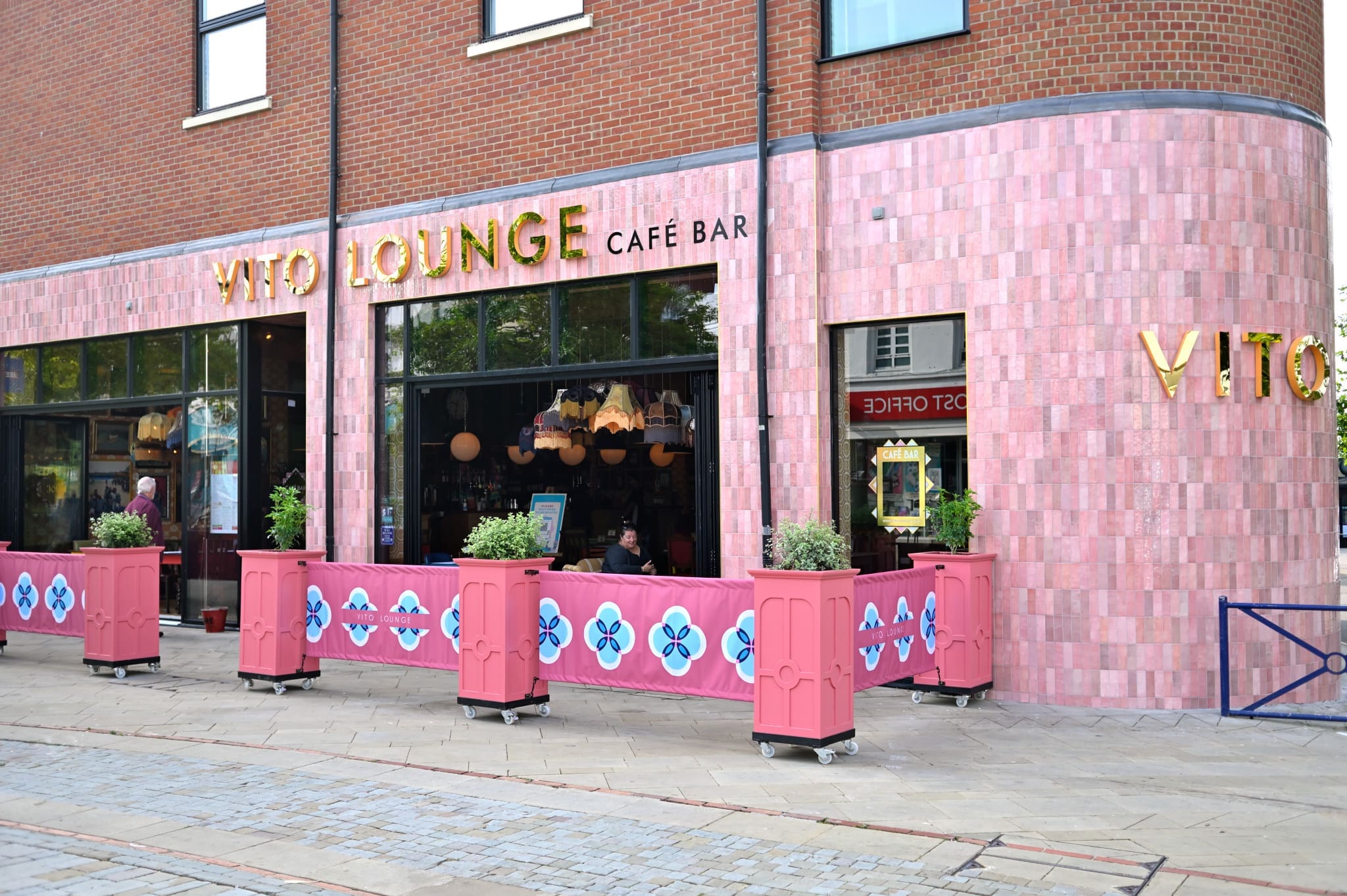 The exterior of Vito Lounge Café Bar features pink tiled walls, large windows, gold Vito signage, and outdoor seating with pink planters along the sidewalk. A few people are visible inside and outside the café.