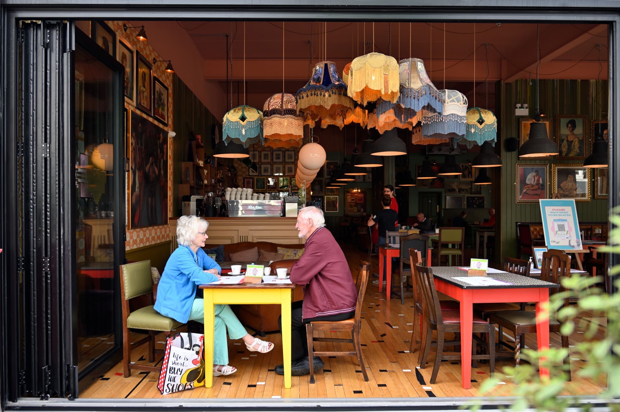 An older couple sits at a brightly colored table inside Vito’s cozy, eclectic café with vintage decor and hanging lampshades, enjoying drinks and conversation. The scene is viewed from outside through a large open window.