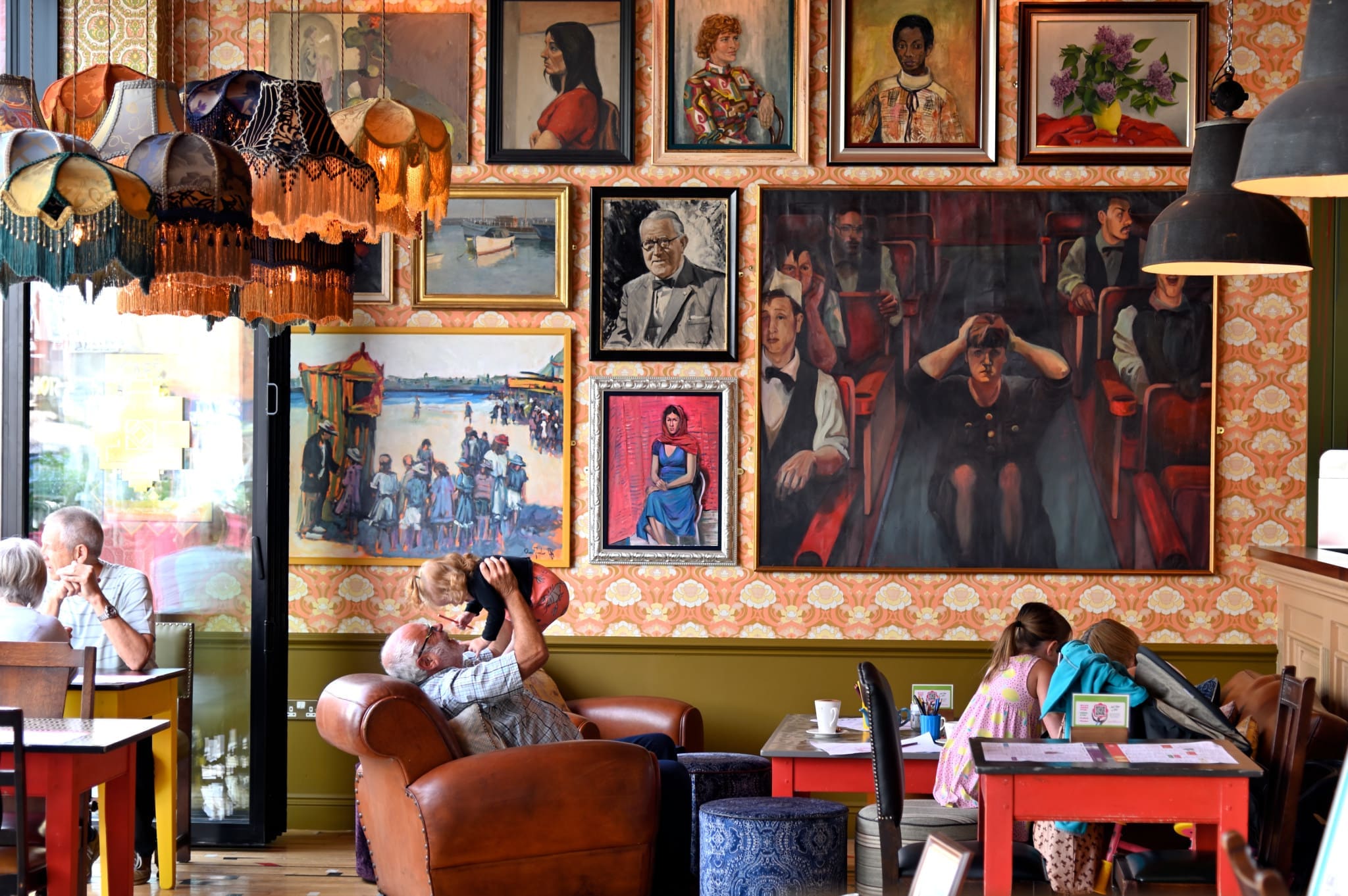 A cozy café with eclectic lamps, vibrant wallpaper, and walls filled with colorful portraits and paintings. People sit at tables and armchairs—perhaps even Vito himself—enjoying drinks and conversation in the warm, artistic atmosphere.