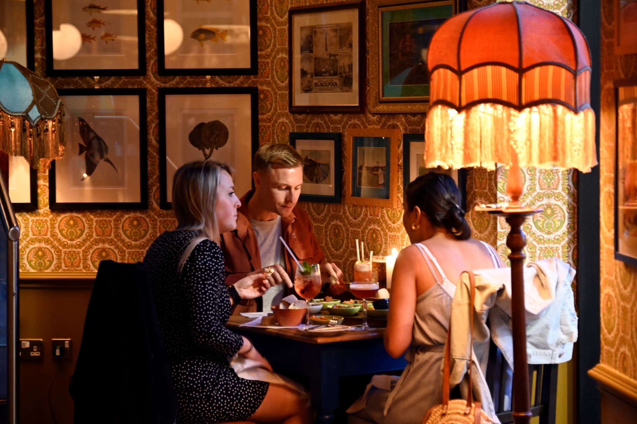 Three people sit around a small table in a warmly lit restaurant, enjoying food and drinks. The walls are decorated with framed art and patterned wallpaper; a vintage lamp adds a cozy, Vito-inspired atmosphere.