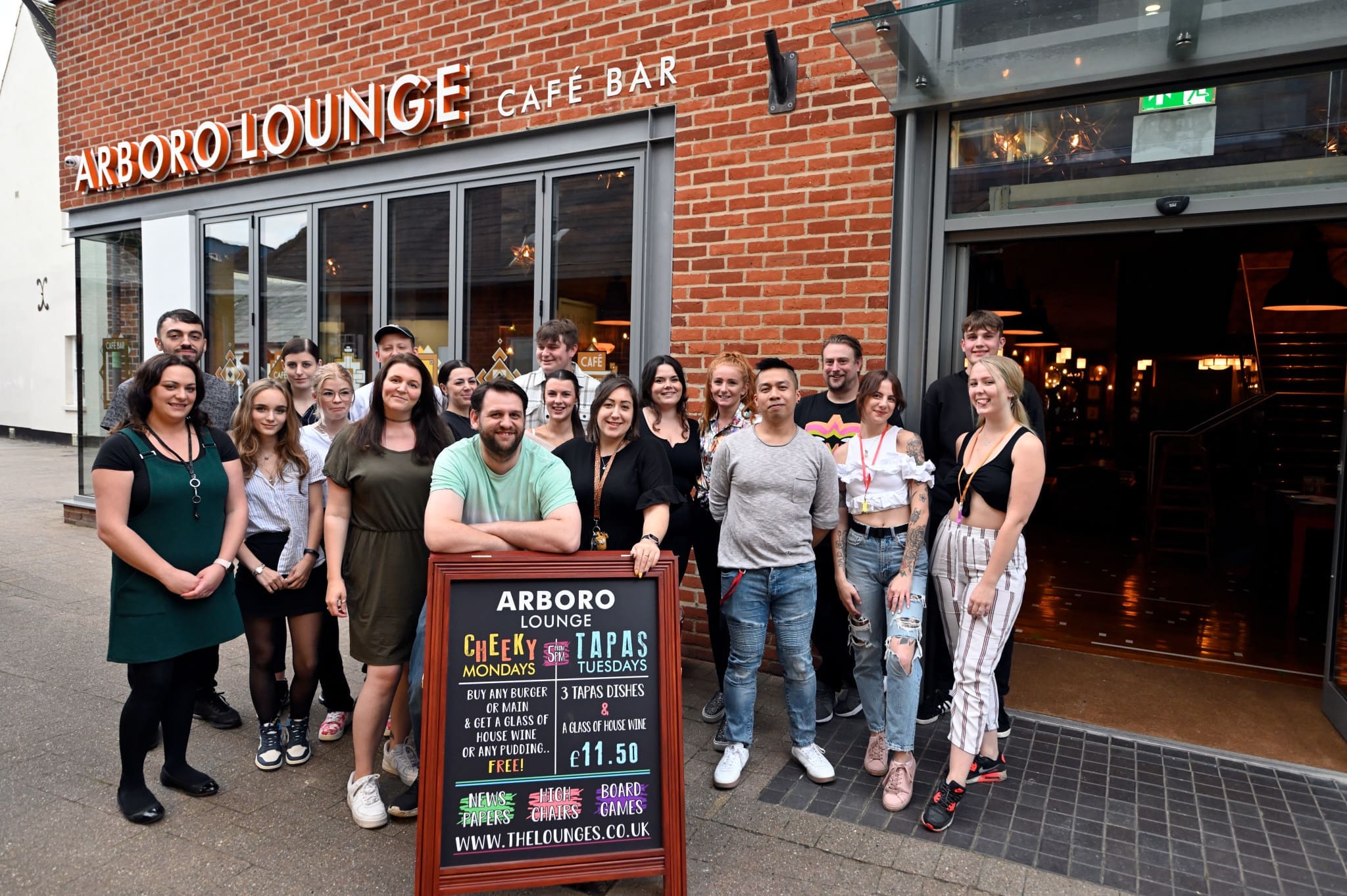 A group of smiling people stand together outside Arboro Lounge café bar, posing behind a menu board on the pavement. The welcoming Arboro entrance and sign are visible in the background.