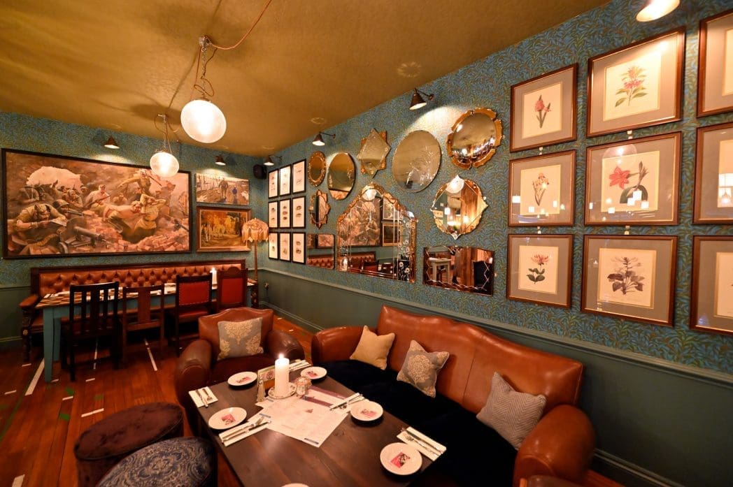 A cozy Arboro restaurant interior with brown leather sofas, a dark wood table set with plates and menus, floral and mirror wall decor, framed art, and warm ambient lighting.