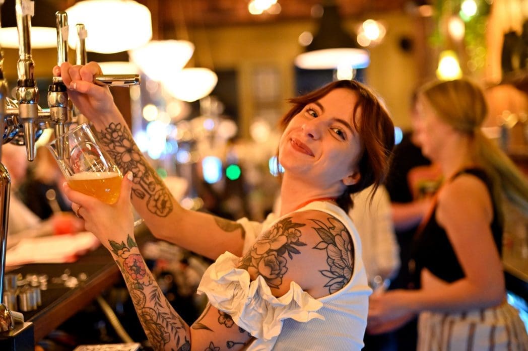 A smiling Arboro bartender with floral tattoos pours a draft beer into a glass at a lively, warmly lit bar. Other people are blurred in the background, adding to the vibrant atmosphere.