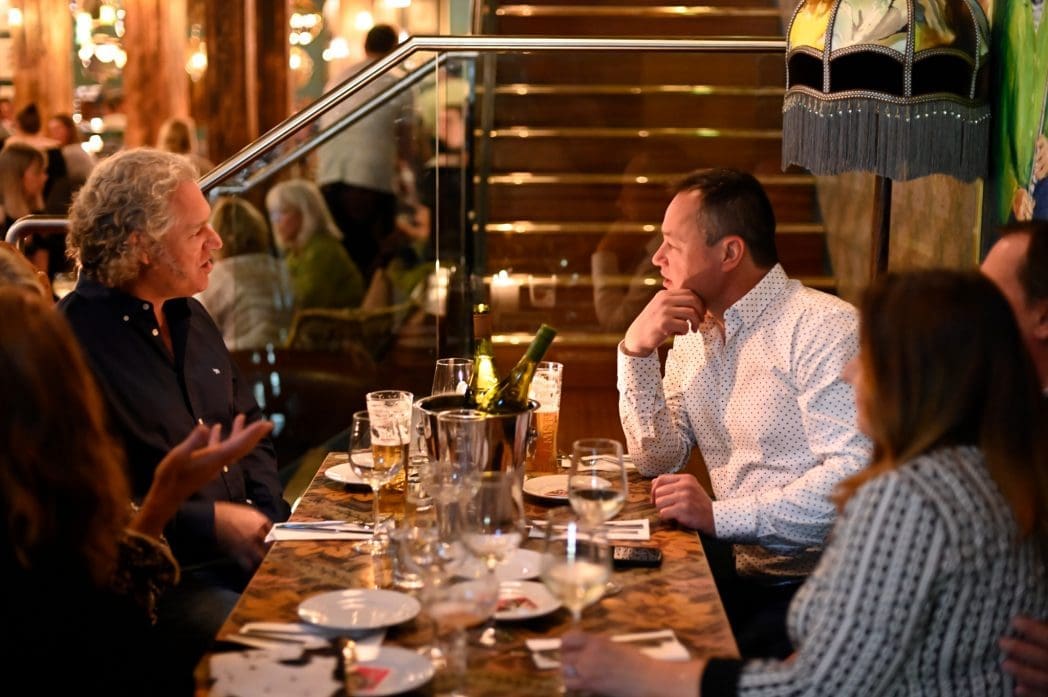 Four people sit around an Arboro restaurant table with drinks, wine glasses, and a wine bucket, engaged in conversation. The warmly lit setting features reflections and other guests in the background.