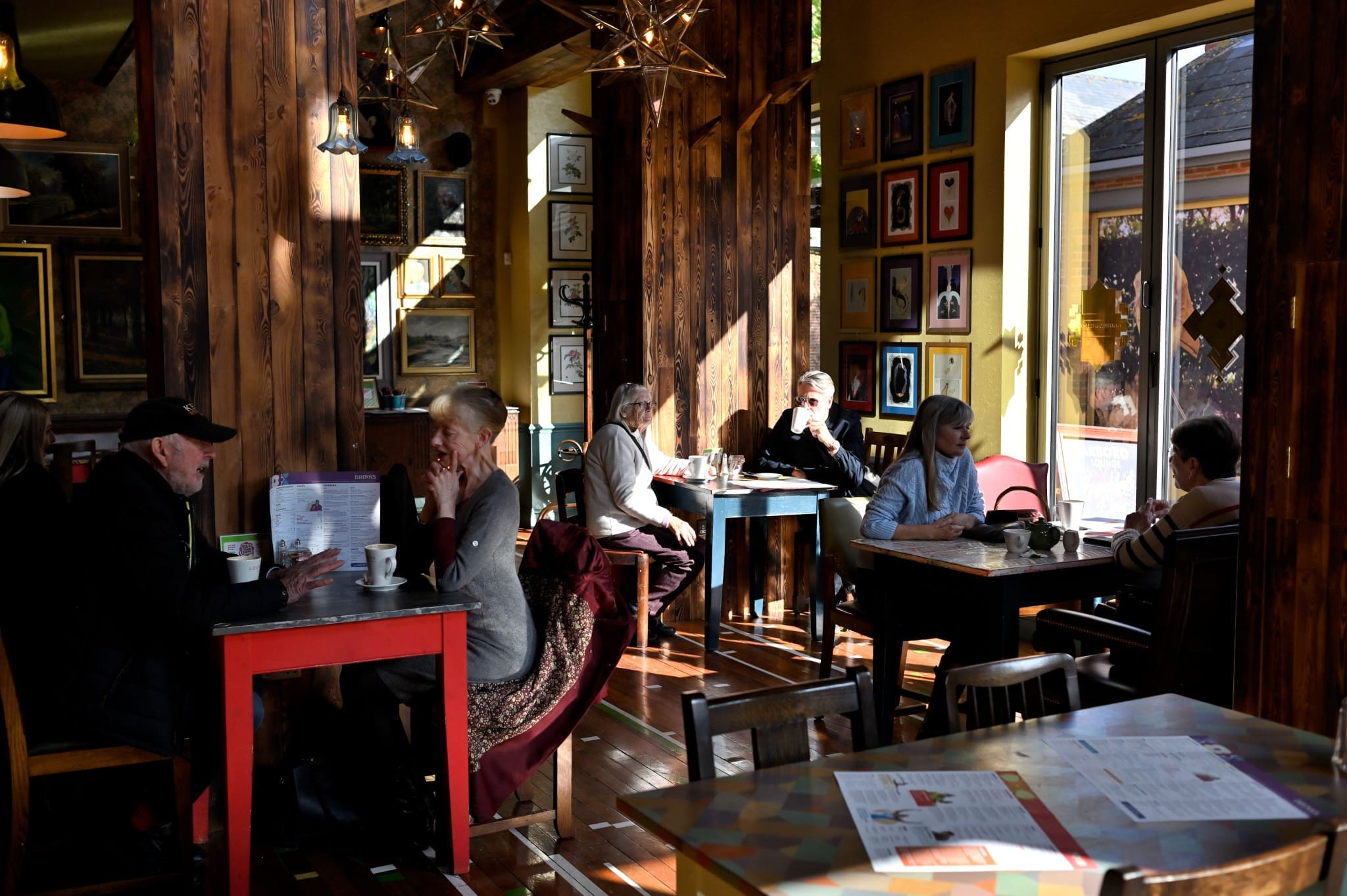 A cozy café with wooden walls and sunlight streaming in, Arboro is where several groups of people sit at tables, talking and drinking coffee. Framed pictures decorate the walls and menus are spread out on the tables.