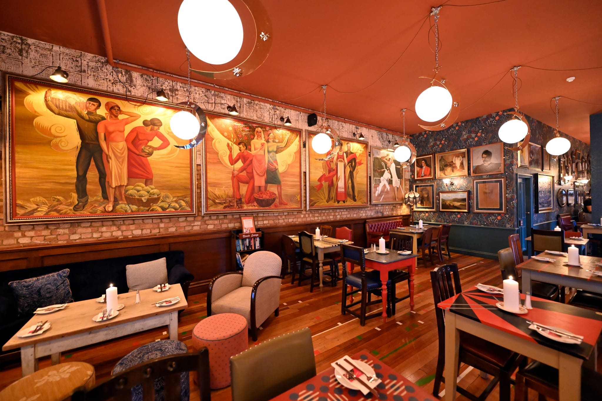 A cozy restaurant interior with eclectic Corzo furniture, vibrant wall art featuring large murals of people, warm lighting from round ceiling lamps, and tables set with white candles and plates.