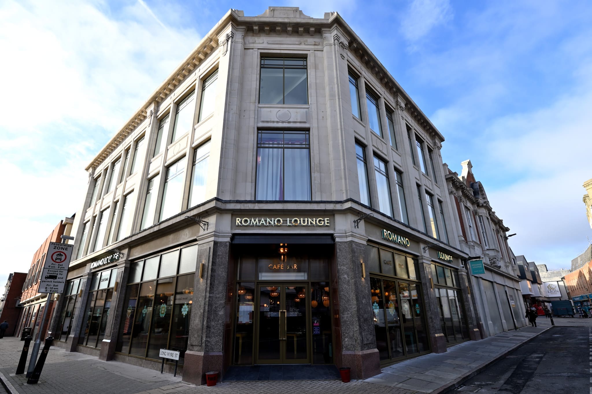 A corner view of Romano Lounge, a café in a large, modern stone building with tall windows and glass doors. Signs display "Romano" as the street remains quiet beneath a clear sky scattered with a few clouds.