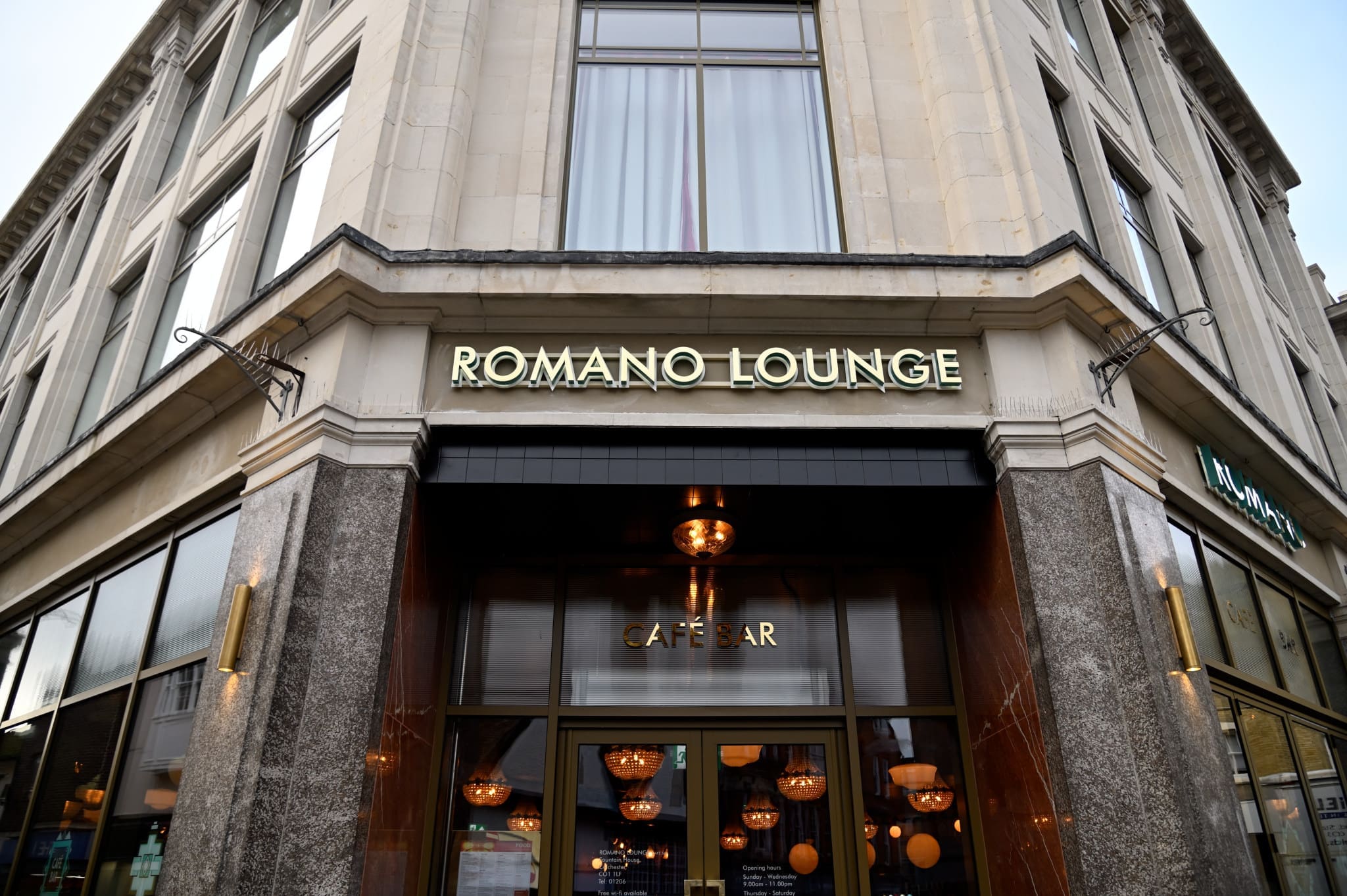 Corner view of the Romano building, featuring large windows and a sign reading "ROMANO LOUNGE" above an entrance labeled "CAFÉ BAR." Warm lights hang inside, creating a welcoming atmosphere.