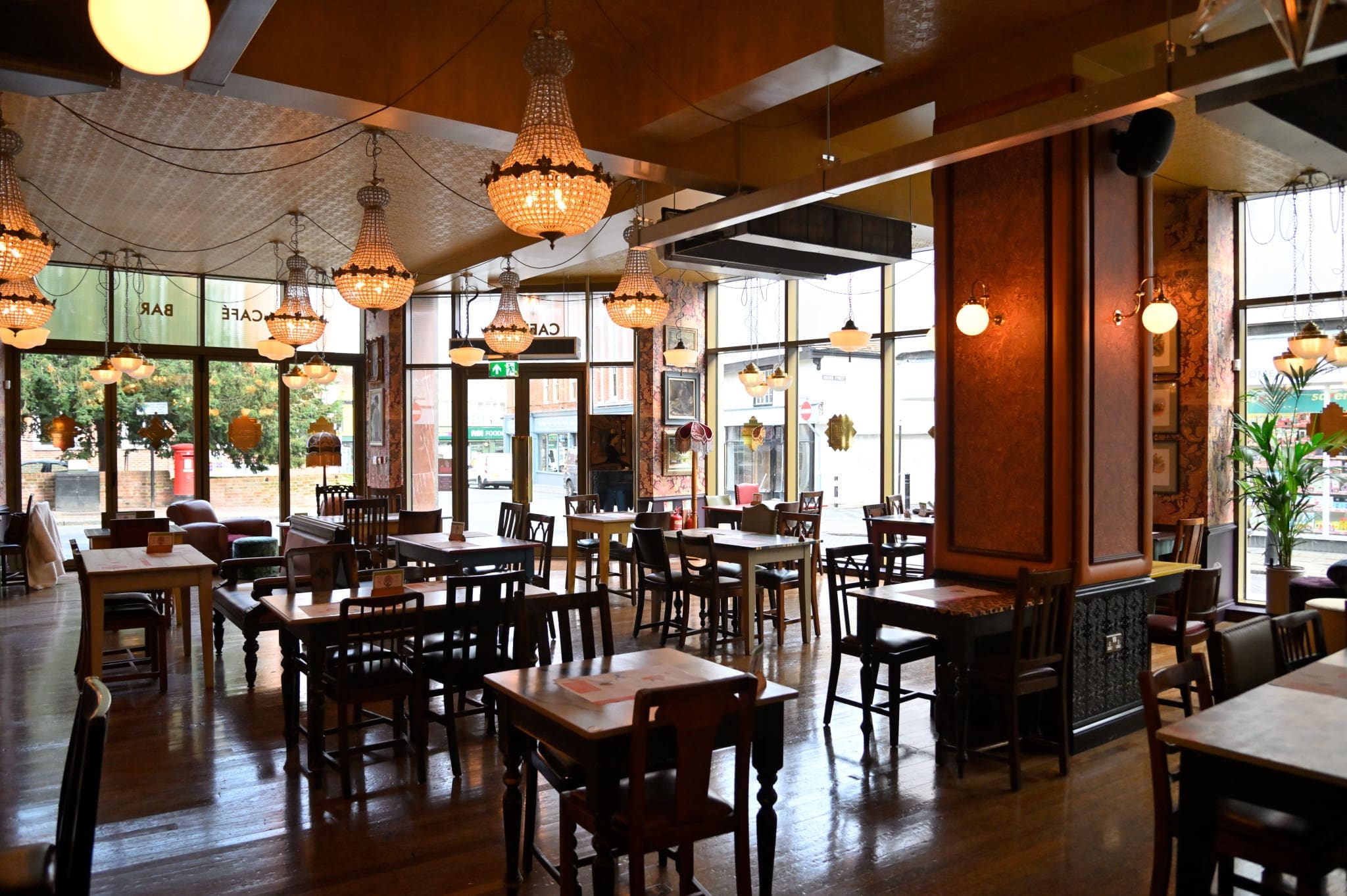 A spacious, warmly lit café with wooden tables and chairs, large windows letting in natural light, ornate Romano chandeliers, and vintage décor. The café appears empty and inviting.