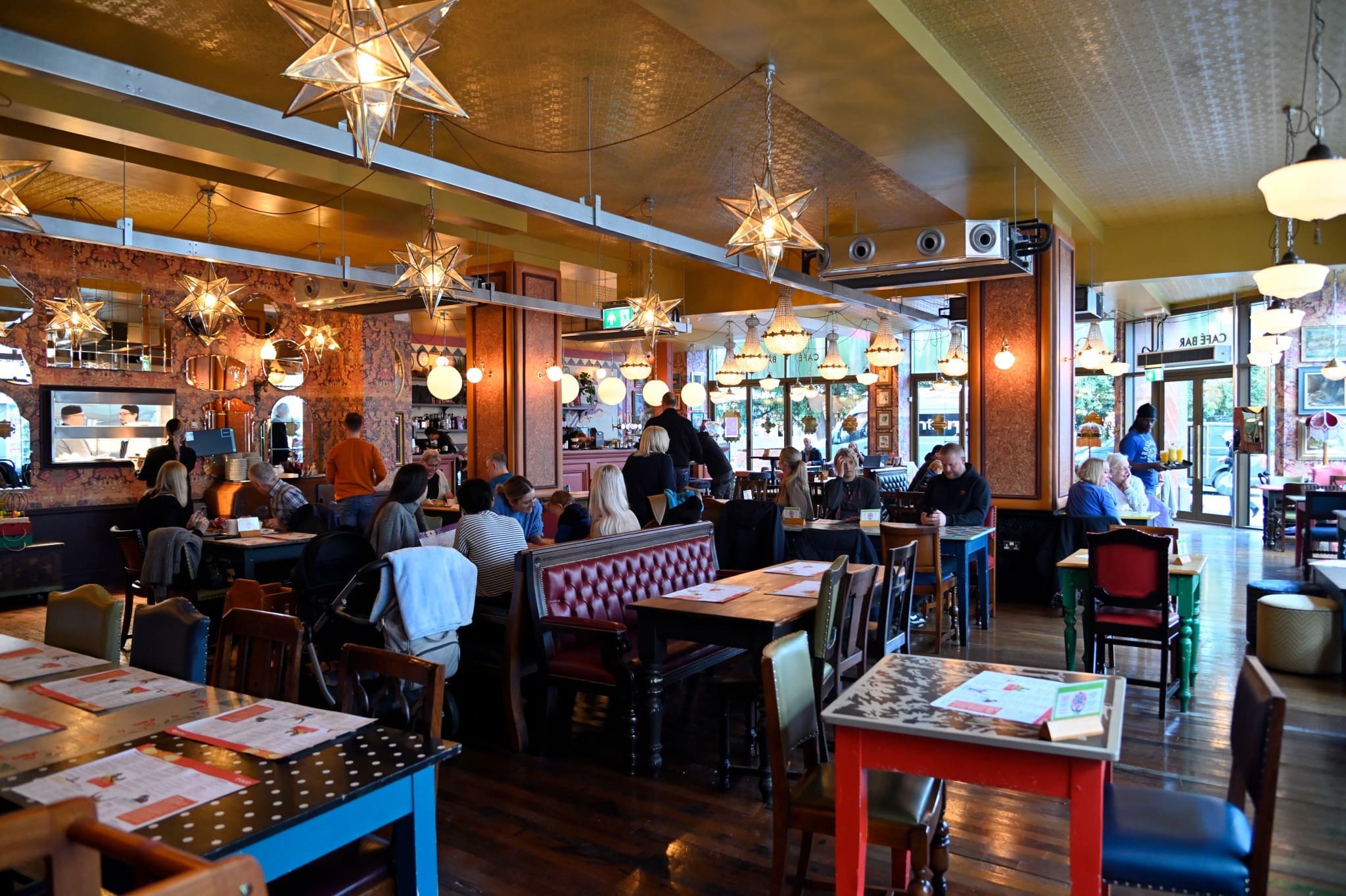 A colorful, lively restaurant interior with eclectic furniture, star-shaped light fixtures, and several groups of people dining and chatting. Large windows let in natural light as menus featuring Romano dishes are placed on the tables.