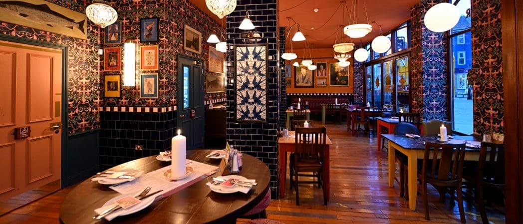 A warmly lit, eclectic restaurant with wooden floors, round and square tables, patterned wallpaper, framed pictures on the walls, blue tiled accents inspired by Bonito style, and globe pendant lights hanging from the ceiling.