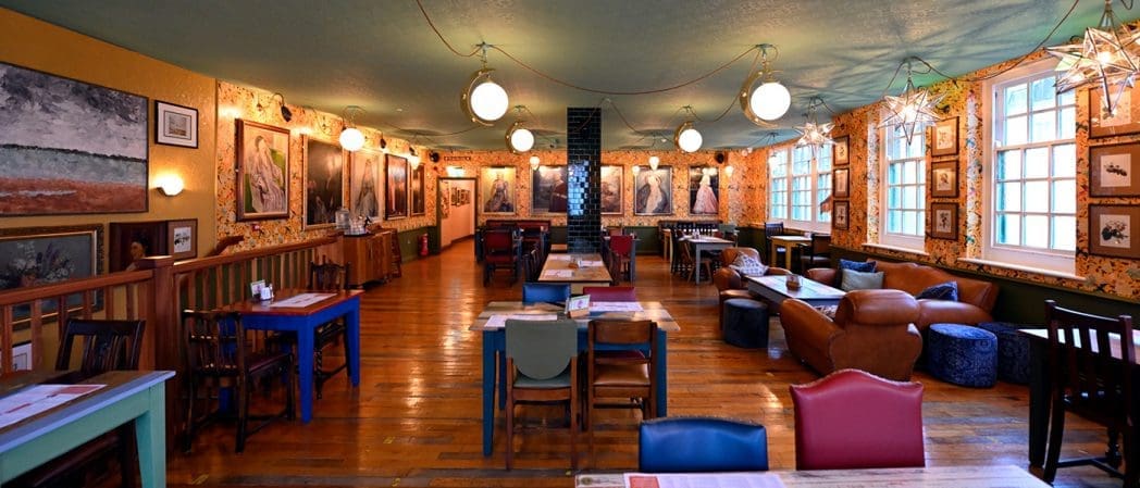 A cozy, eclectic café interior with colorful chairs and wooden tables, vibrant Bonito wallpaper, paintings and photos on the walls, star-shaped pendant lights, and large windows letting in natural light.