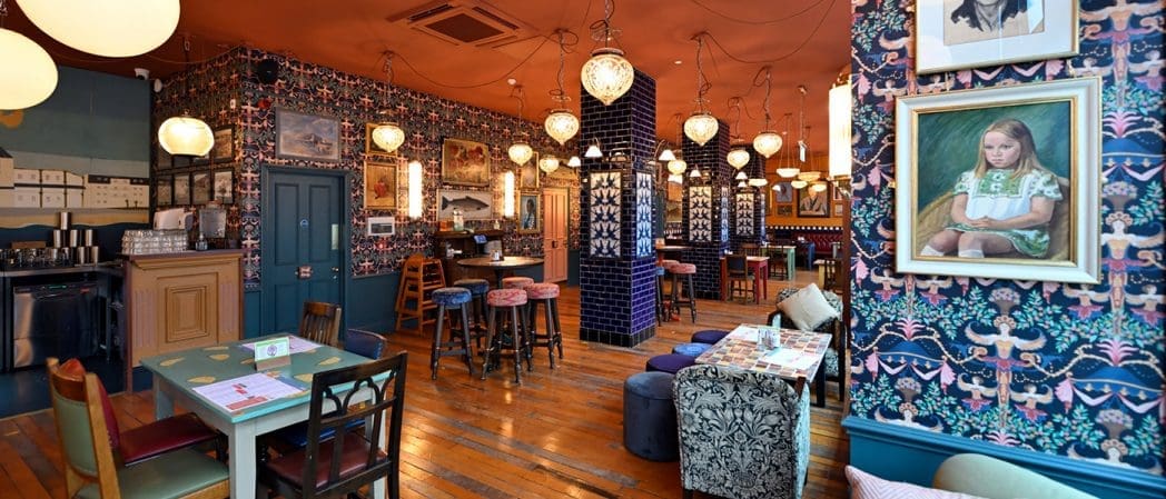 A cozy, eclectic cafe interior with patterned wallpaper, hanging lamps, mismatched furniture, wooden floors, and framed art—including a Bonito portrait of a girl in a green dress on the right wall.