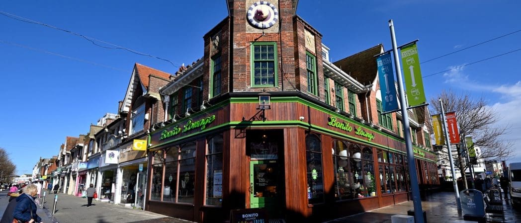 Corner view of a busy shopping street with a two-story brick building featuring green trim and the Bonito Lounge restaurant. People stroll under a clear blue sky, passing colorful banners and vibrant shopfronts.