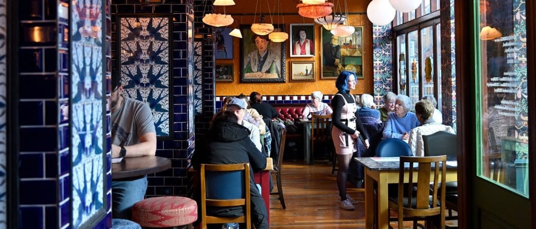 A vibrant café with colorful tiled walls and framed portraits, where people are dining and chatting at wooden tables. A server with blue hair stands by a group, and natural light streams in through large windows gives the space a bonito charm.
