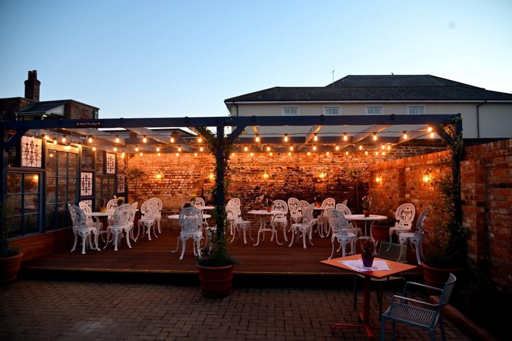 An outdoor patio at Correo features white ornate metal chairs and tables under string lights, surrounded by brick walls and potted plants, creating a cozy evening atmosphere. A single table stands in the foreground.