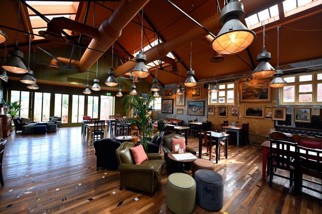 Spacious, sunlit café with wooden floors, eclectic furniture, and large windows. The inviting Correo vibe comes through framed art on the walls and lush plants, while hanging industrial lights complete the cozy atmosphere.