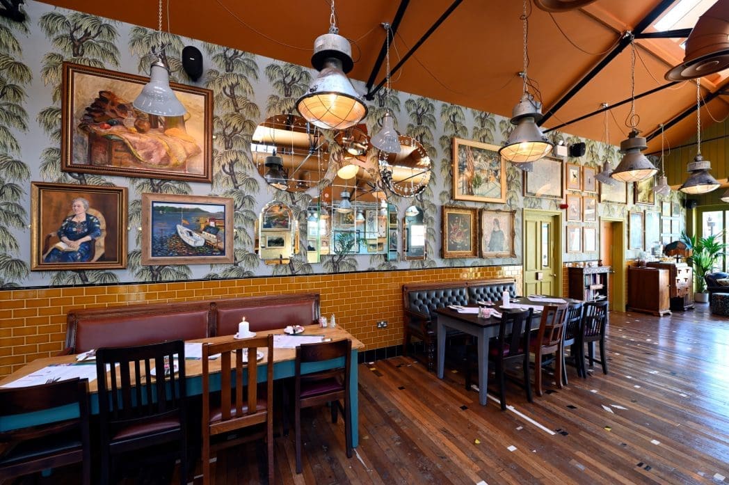 A stylish restaurant interior with wooden floors, Correo-inspired artwork and mirrors on patterned wallpaper, a mix of wooden tables and leather seating, industrial light fixtures, and a warm, inviting atmosphere.