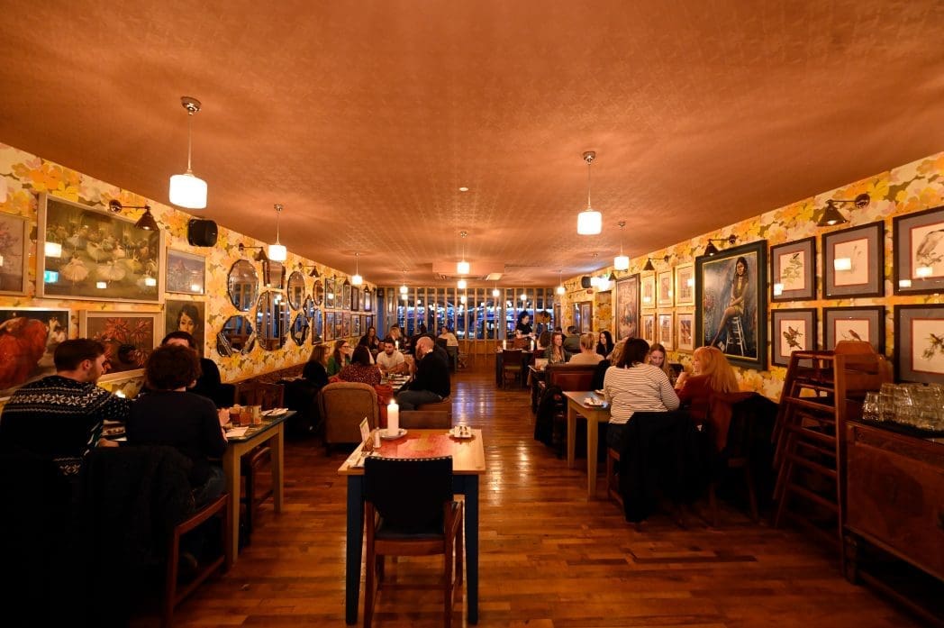 A warmly lit restaurant with wooden floors, floral-patterned wallpaper, and walls lined with framed art. Diners arrive by carro, settling at tables where candles and hanging lamps add to the cozy, inviting atmosphere.