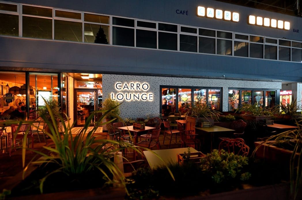 Outdoor view of Carro Lounge café and bar at night, with illuminated Carro signs, potted plants, and empty tables on a patio. Inside, warm lighting and people are visible through large windows.
