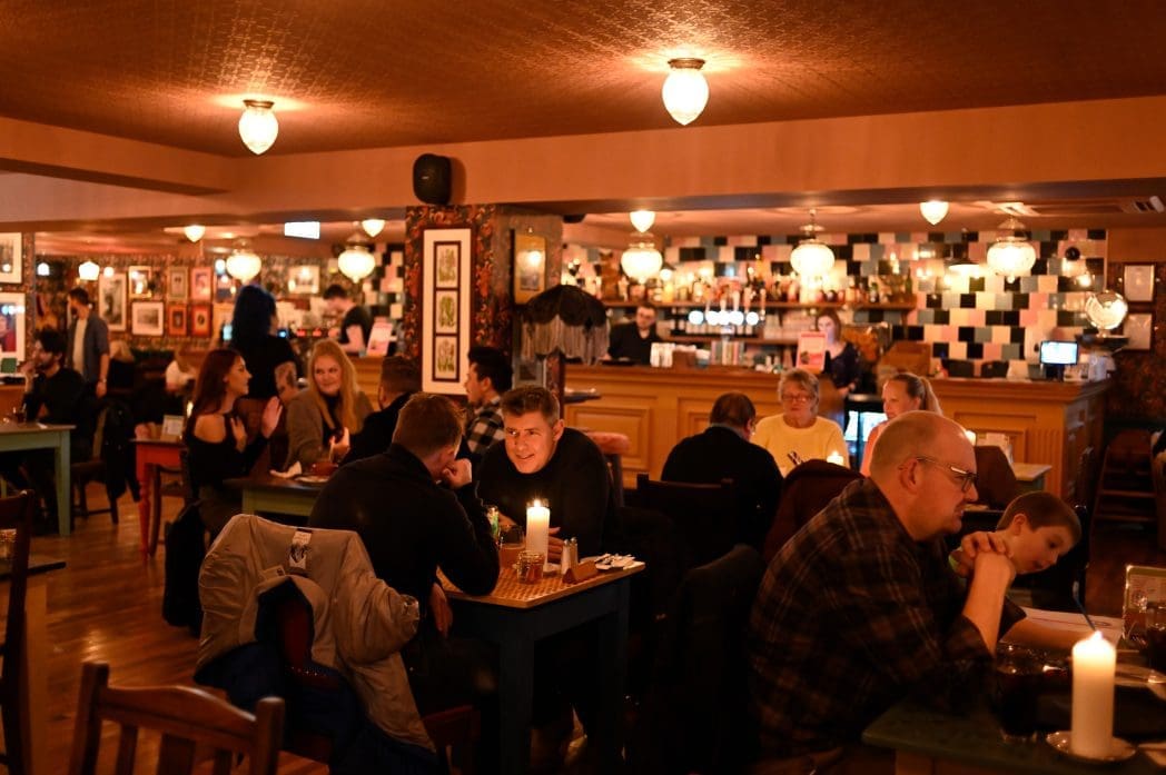 A warmly lit restaurant with people seated at tables, eating and talking. Some guests chat while others focus on their meals, and a Carro-themed decorative wall art adds charm to the bar area visible in the background.