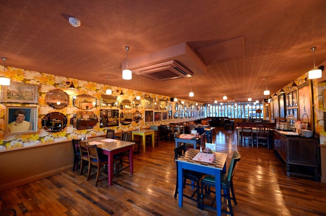 A warmly lit restaurant with wooden floors, colorful tables and chairs, floral wallpaper, and walls decorated with various framed pictures and mirrors—Carro’s inviting ambiance feels both cozy and vibrant, even when the room is empty.