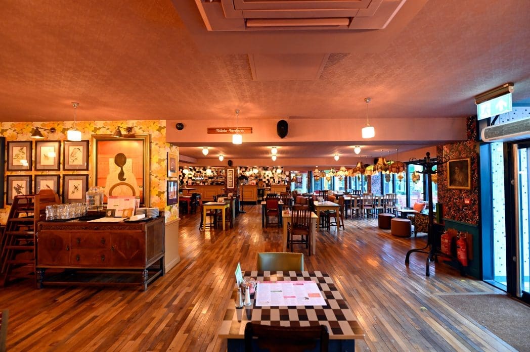 Spacious Carro restaurant interior with wooden floors, eclectic wall art, a mix of tables and chairs, soft lighting, and large windows filling the space with natural light. The warm, inviting atmosphere is enhanced by decorative touches.