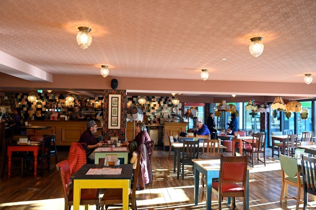 A brightly lit café with colorful chairs and wooden tables, patterned ceiling, decorative hanging lamps, and people dining and talking. Through the large windows, a parked carro is visible, adding charm to the warm, inviting atmosphere.