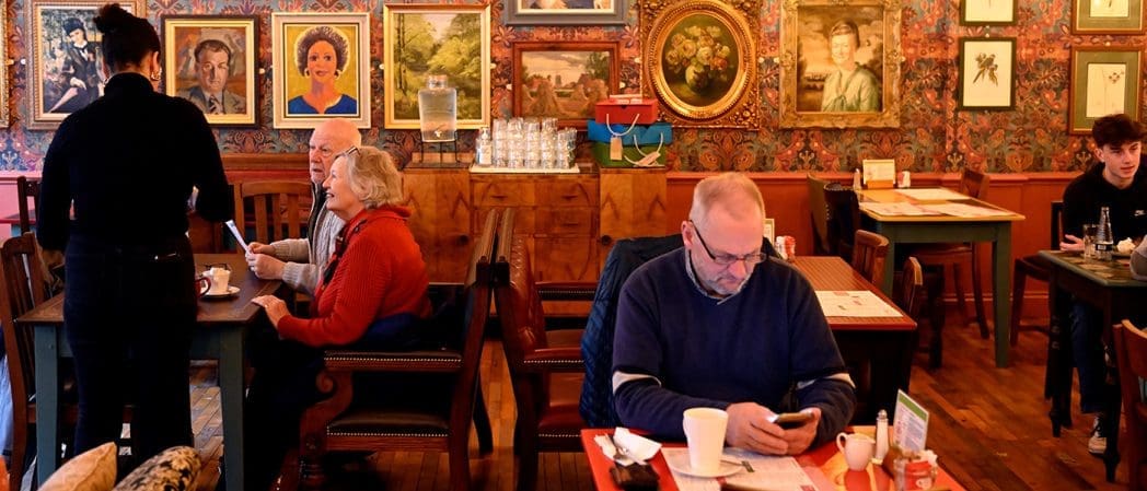 A cozy café with several people seated at tables. A waitress takes an order from an older couple, while a man at the front table looks at his phone. Clavio's portrait paintings and framed art cover the wallpapered walls.