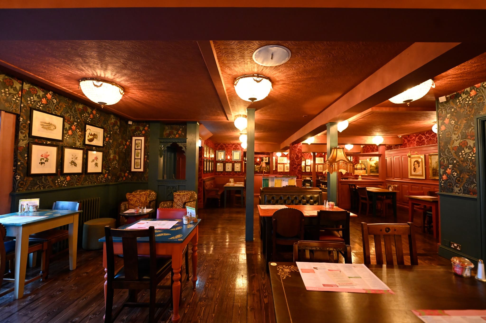 A cozy, warmly lit restaurant with vintage decor, patterned wallpaper, wooden floors, and framed artwork on the walls—Hudo’s inviting space features various wooden tables and chairs spread throughout the room.