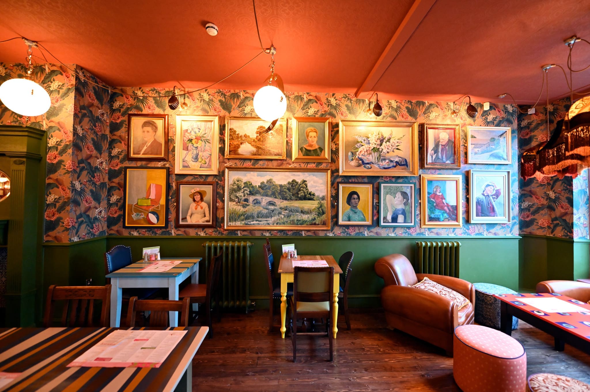A cozy, colorful cafe interior with floral wallpaper, green walls, and a gallery wall of framed portraits—this Hudo-inspired space features eclectic furniture including tables and armchairs, creating an inviting and artistic atmosphere.