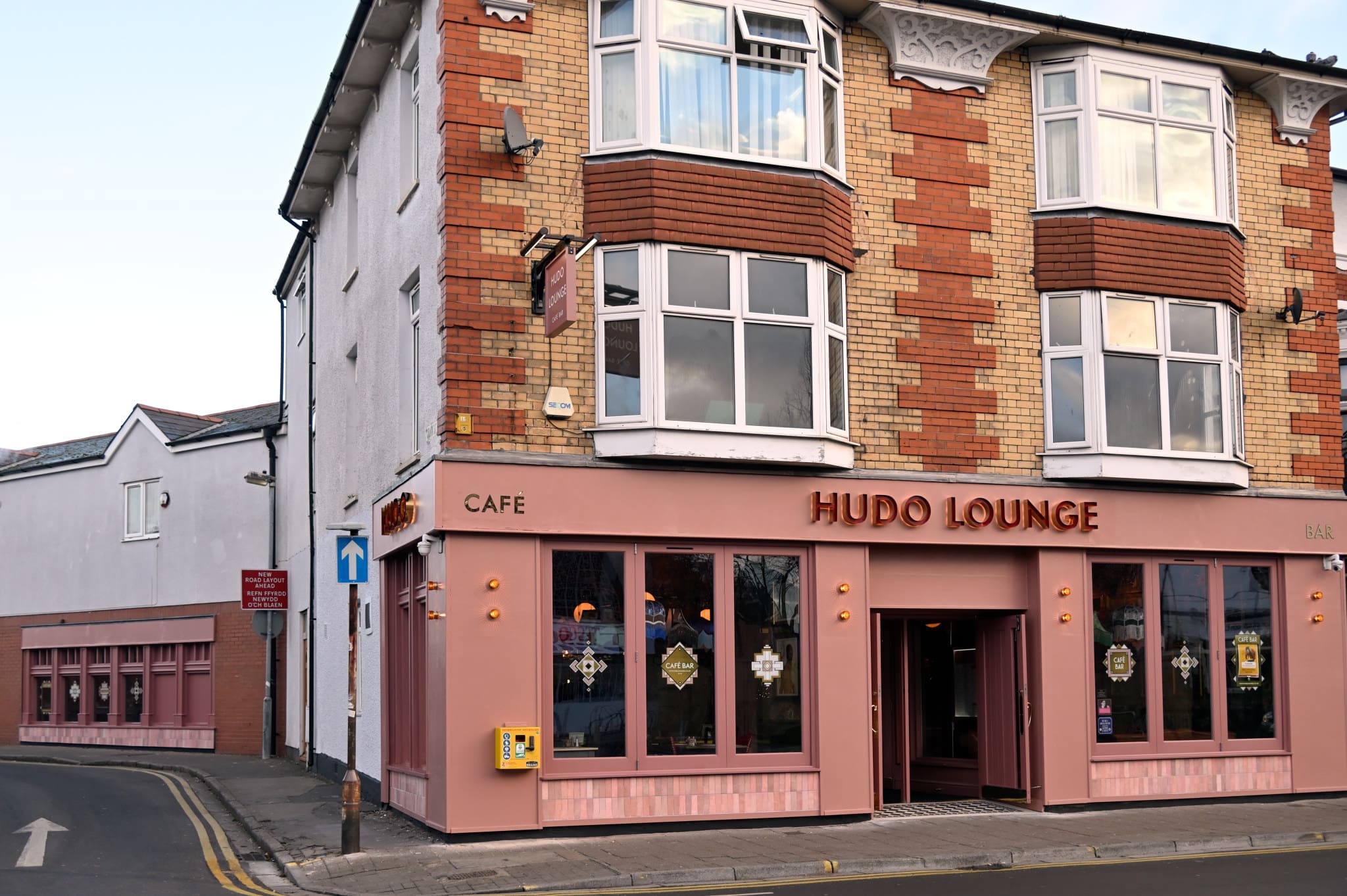 A street corner view of Hudo Lounge, a café and bar in a three-story brick building with large windows and pink accents, located at the junction of two roads—a one-way street sign points toward Hudo’s entrance.