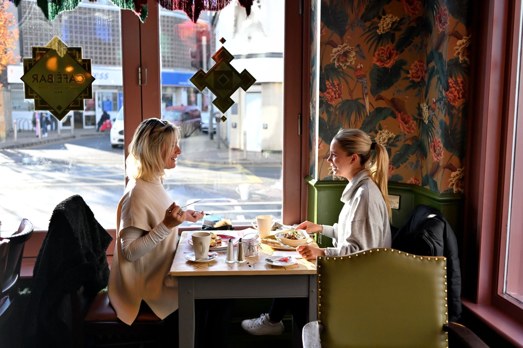 Two women sit across from each other at a table in Hudo, a cozy cafe with floral wallpaper and warm decor. Sunlight streams through the window, illuminating their cheerful conversation as they enjoy delicious food and coffee.