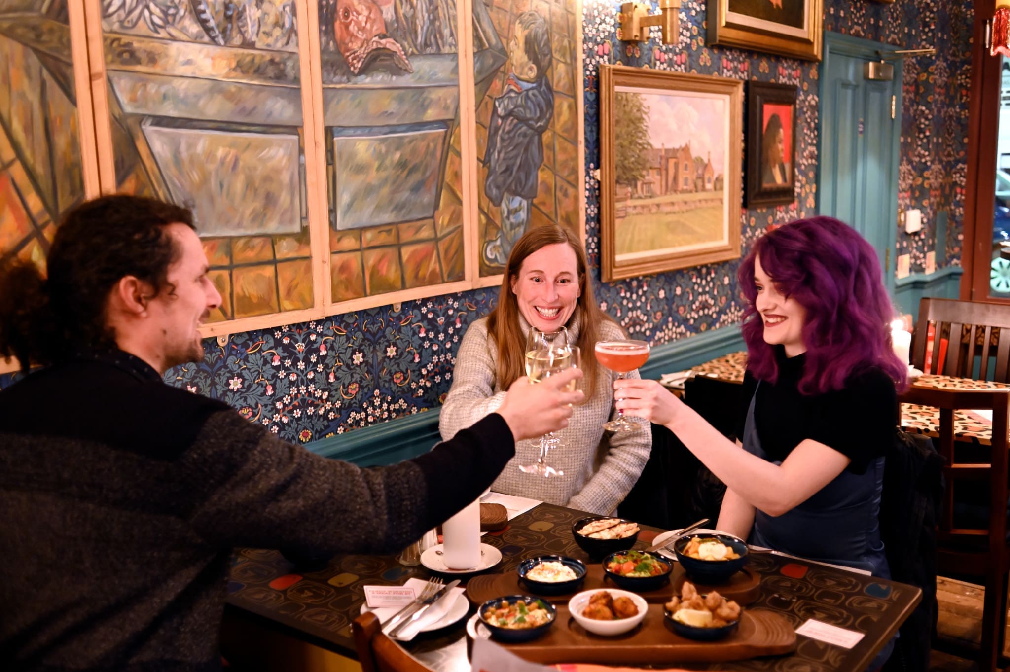 Three people sit at a Cortillo restaurant table, smiling and raising glasses in a toast. The table overflows with delicious dishes, while vibrant artwork and patterned wallpaper warm the cozy, inviting room.