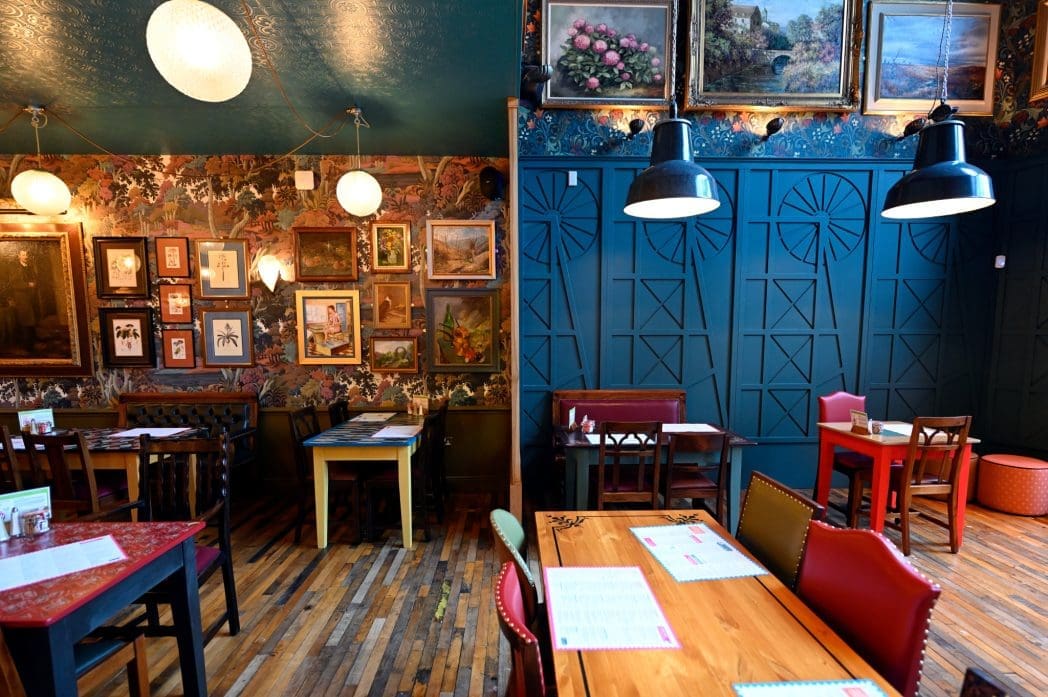 A cozy Cadno restaurant interior with eclectic decor; one side has floral wallpaper, framed paintings, and round lanterns, while the other features teal walls, geometric patterns, and hanging black lamps over wood tables.